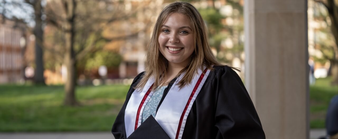 EKU student, Lexie Barth, wears her graduation gown and holds her cap outside of the Combs building on EKU's campus.