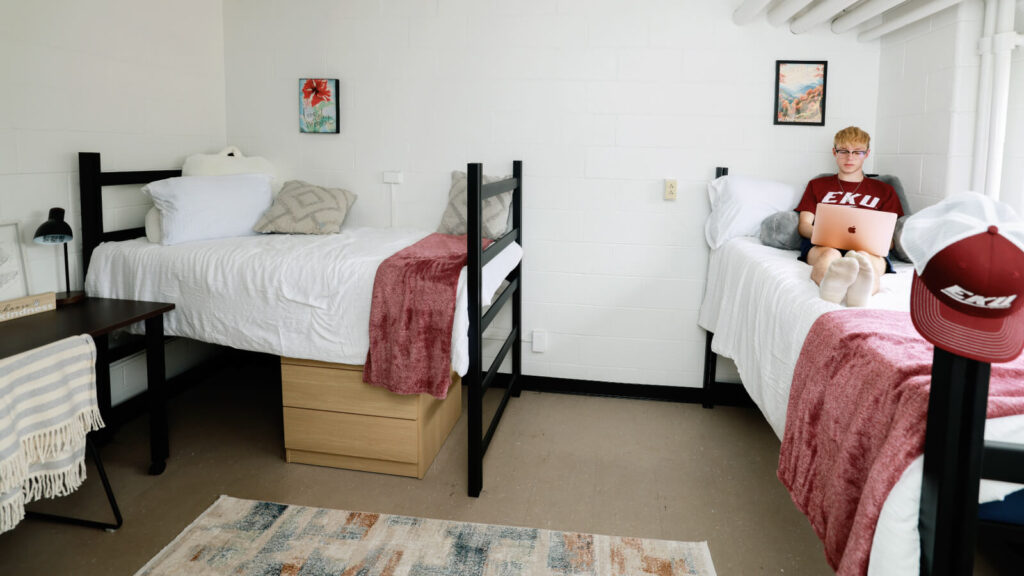 A student uses a laptop on one of two lofted beds with storage underneath in Mattox Hall. One of two study tables is in view.