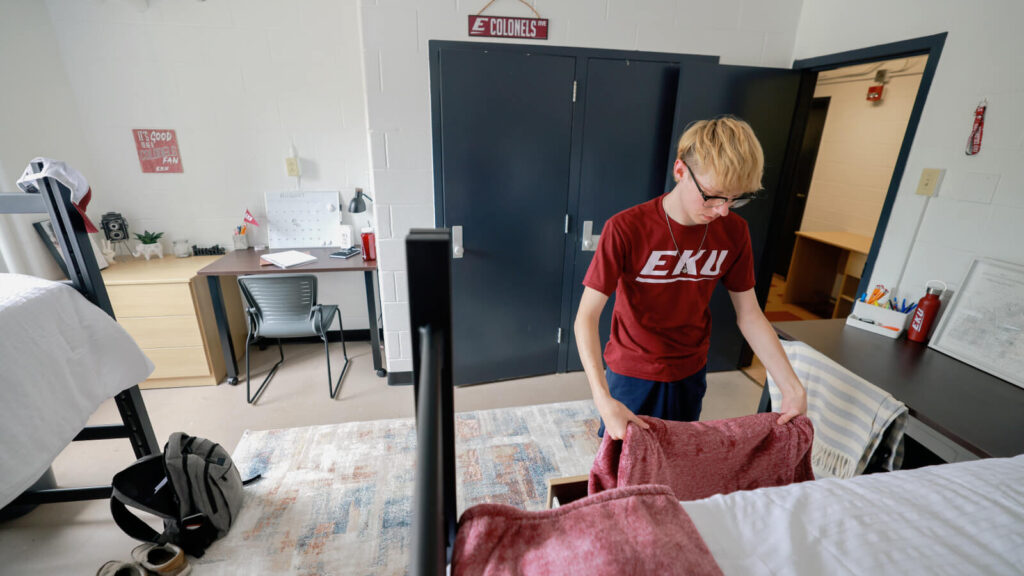 a student puts a blanket in a drawer under a lofted bed across from a study table and closet in a shared room in Mattox Hall