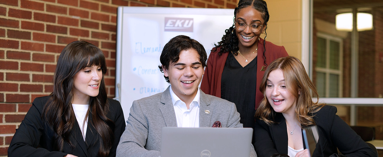 Professionally dressed EKU female business students look on as male EKU student works on laptop in the business technology center