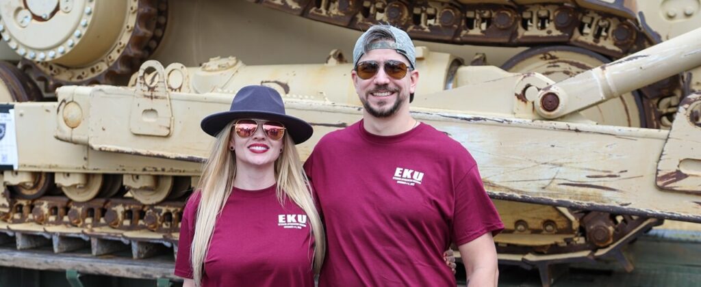 April Bixler, ’14, Army veteran and EKU graduate, and her husband wear EKU t-shirts while standing in front of an Army tank.