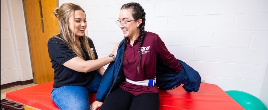 A female occupational therapy student helps another student out of a jacket in an OT room on campus