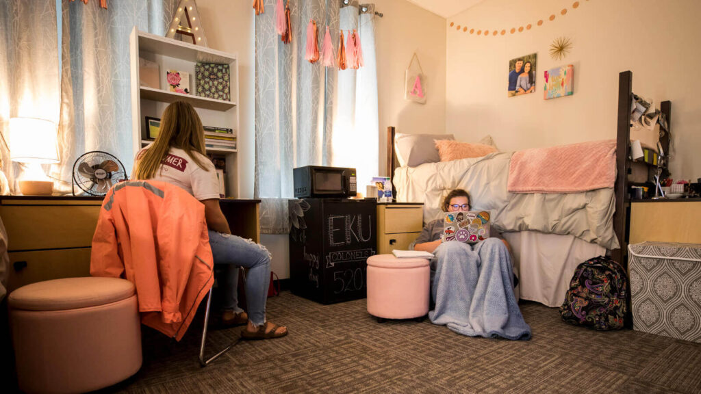 A student sits at a desk while another sits in a chair and uses a laptop by a bed in a shared room in South Hall. A microwave and refrigerator and windows are in view.