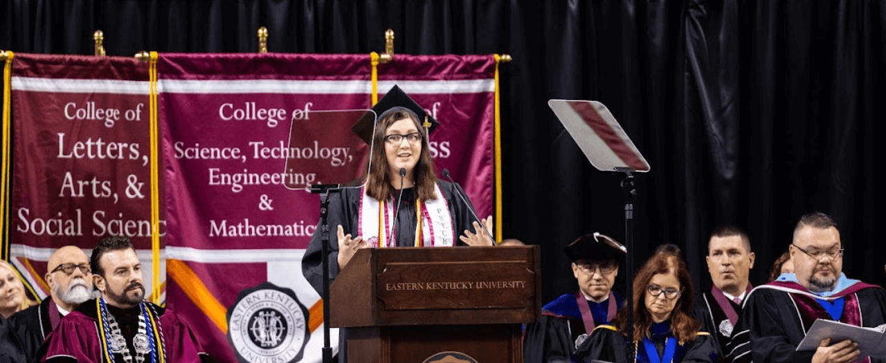 EKU graduate, Holdyn Morrow, delivers the commencement address with EKU faculty and administration seated behind her.