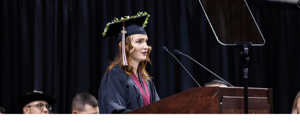 EKU graduate Isabelle Orth in her adorned cap and gown delivers the commencement speech into a microphone at the podium.