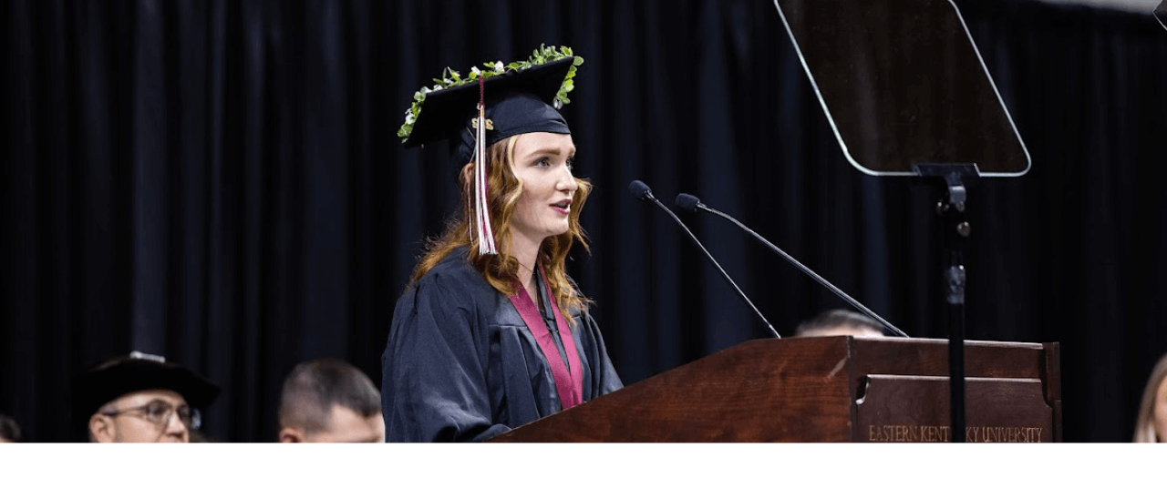 EKU graduate Isabelle Orth in her adorned cap and gown delivers the commencement speech into a microphone at the podium.