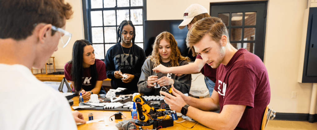 A group of male and female EKU STEM students gather around a table to work on a robotic arm.