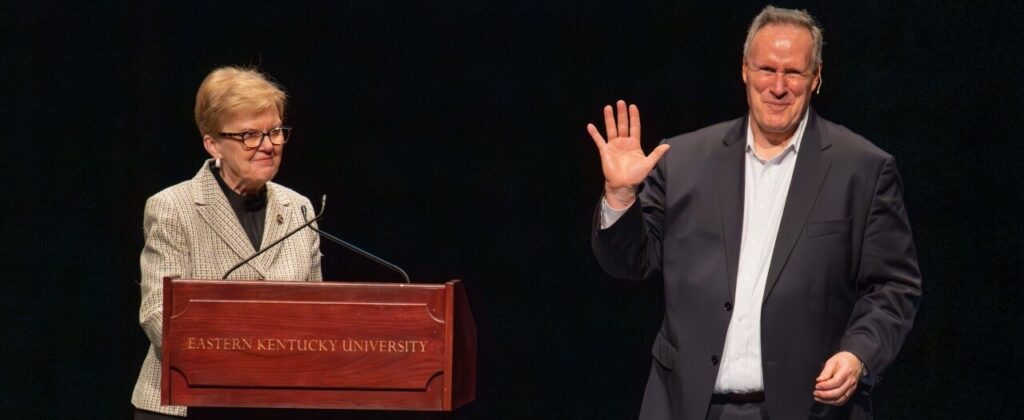 Smiling, Author Stephen M.R. Covey waves to the audience from the stage after Dr. Janna Vance introduces him.