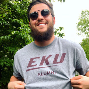 Outside and wearing sunglasses and a beard, Matt Agee pulls out his shirt to display the EKU Alumni logo