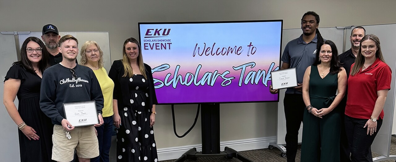 Scholars Tank participants stand next to a large TV displaying a welcome to the event.