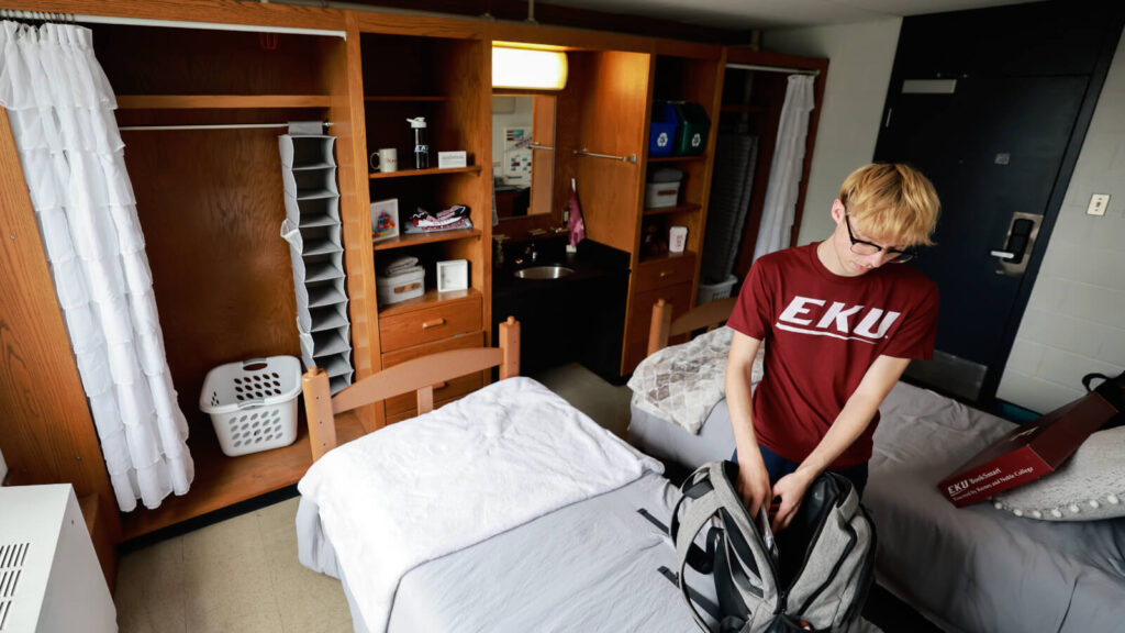 A shared room in Clay Hall has a sink between closets and cabinets. Two beds are in view as a student packs a backpack on one.