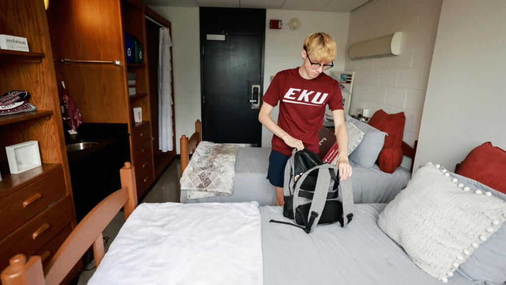 A shared room in Clay Hall has a sink and cabinets and closets and two beds. A student opens a backpack on one bed.