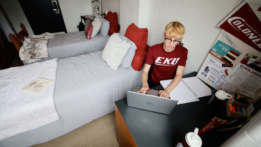 a student sits at a desk in a shared room in Clay Hall with two beds and another desk in view