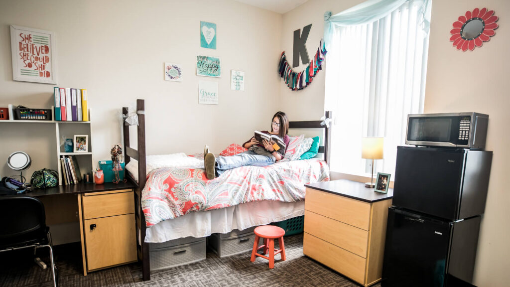 a student sits on a bed in North Hall with storage underneath and a desk and cabinet and small refrigerator and a microwave and windows