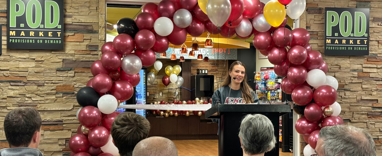 EKU student, Brittany Collins, speaks from a podium in front of a maroon balloon arch in front of the P.O.D. market in Stratton at the ribbon cutting.