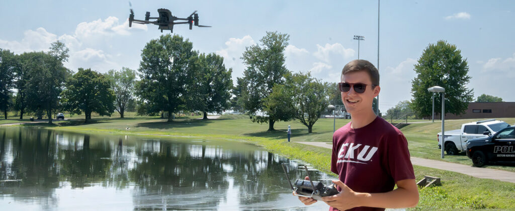 A young male student wearing a maroon EKU t-shirt and sunglasses operates a drone over the pond behind EKU's Stratton building.