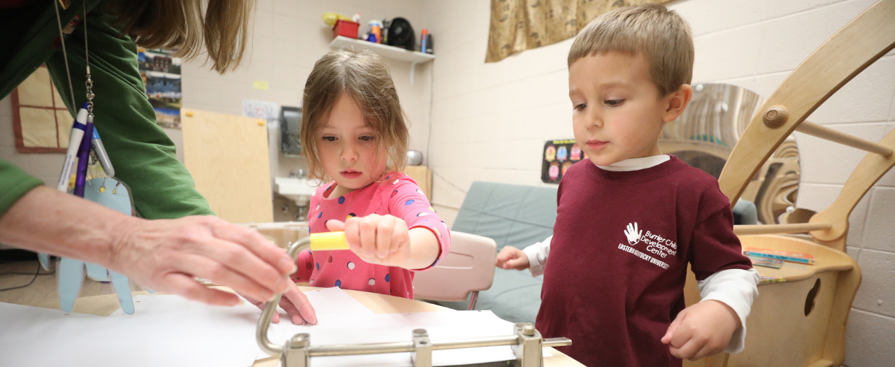 A teacher helps two toddlers use a hole punch for a project they are working on in the Corbin Childcare Center.