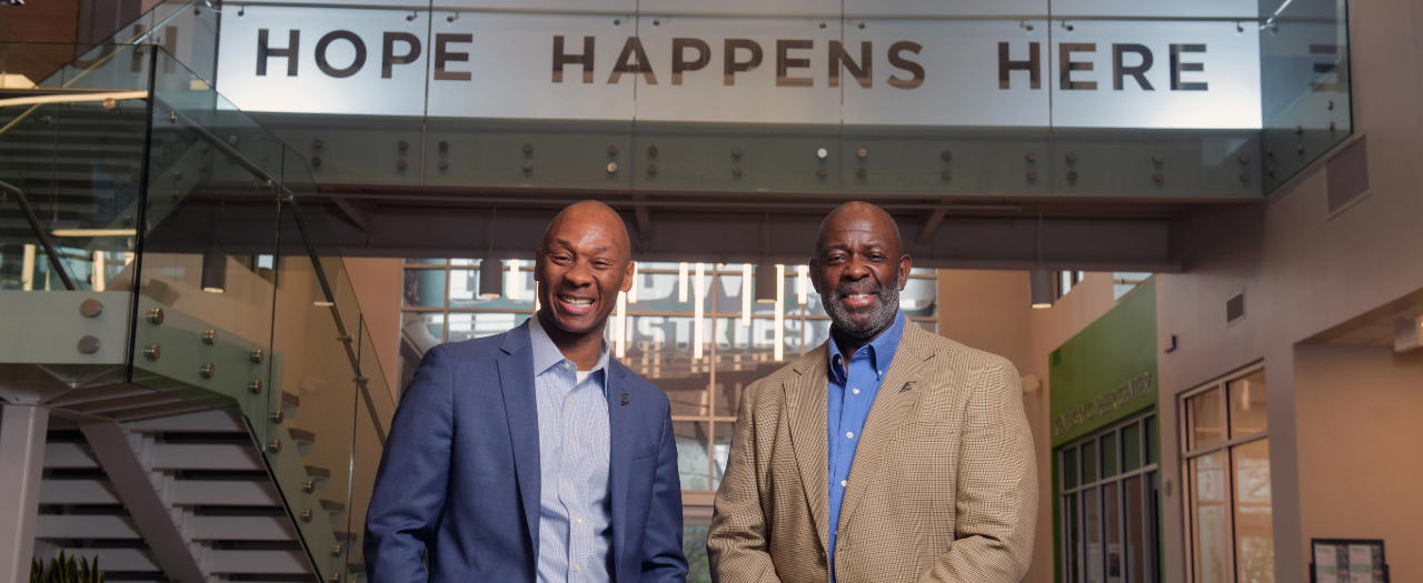 DeVone Holt and Emery Lee, both dressed in suits, smile and stand in front of the Hope Happens Here sign at the Norton Healthcare Goodwill Opportunity Campus.