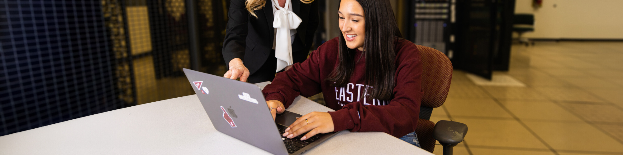 a standing professor points at laptop as a sitting student types on the same laptop in front of a bank of network servers