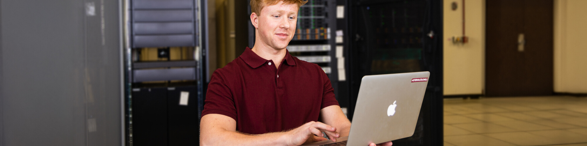 a student uses a laptop while sitting in front of a bank of network servers