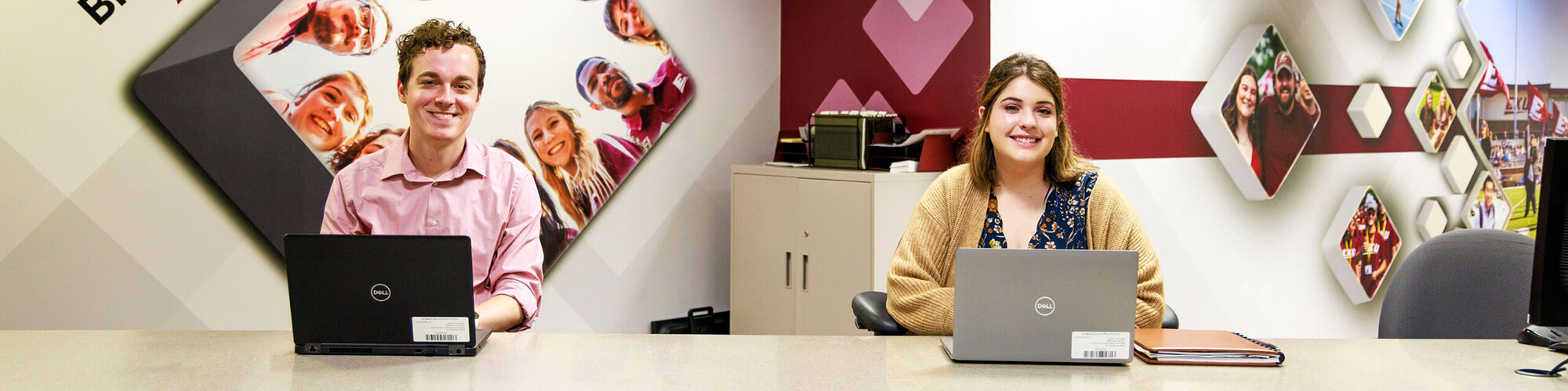 One male and one female EKU employee sit smiling behind computers, ready to help at EKU's Big E Central office.