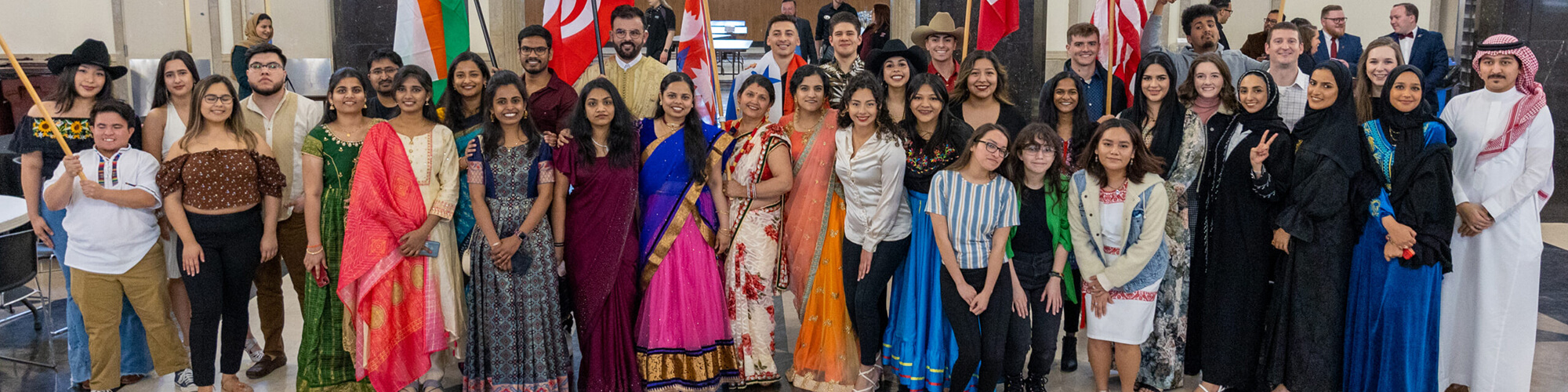 A large group of EKU international students gather in front of flags for a photo, many dressed in attire representing their native countries.