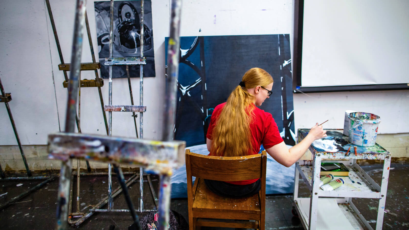 Student in an art studio surrounded by several easels is sitting with their back away from the camera, dips their paint brush into paint on a cart as they are painting on a large canvas that sits in front of them.