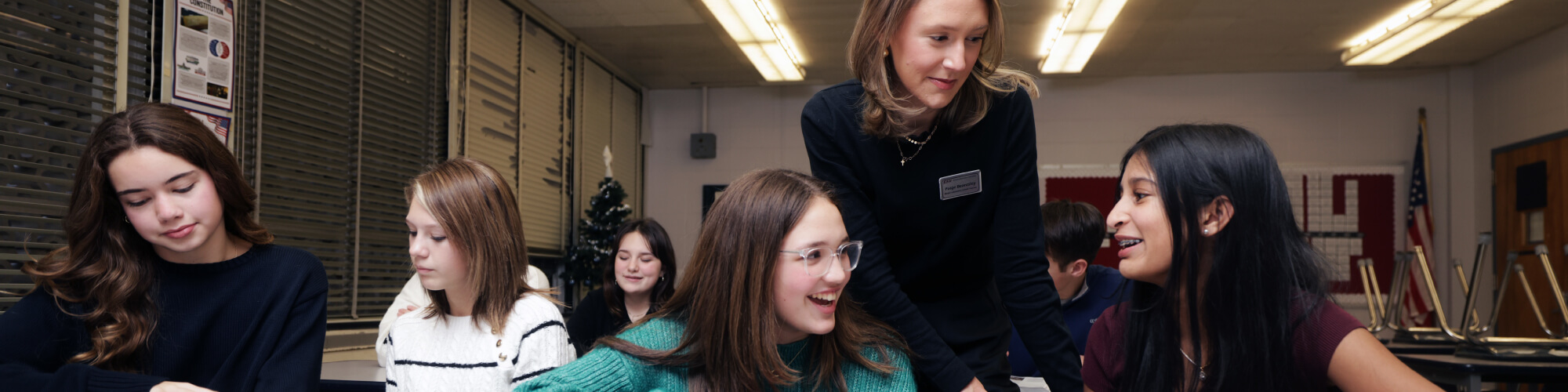 teacher in a classroom setting leans over to help middle school age kids on an assignment at a desk with other students also working on assignments around them