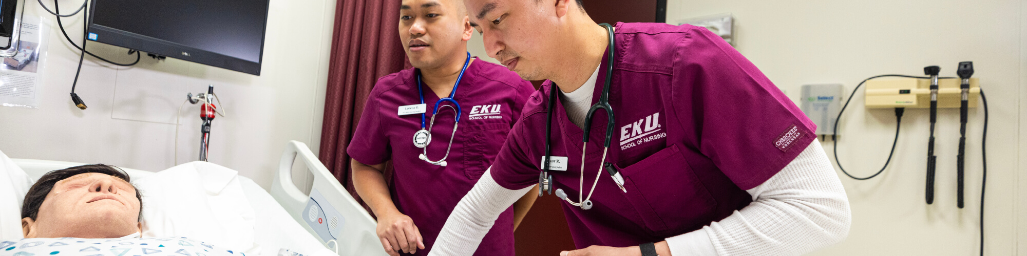 two EKU nursing students wearing maroon scrubs and stethoscopes are practicing checking the pulse on the wrist of a manikin patient in a hospital bed