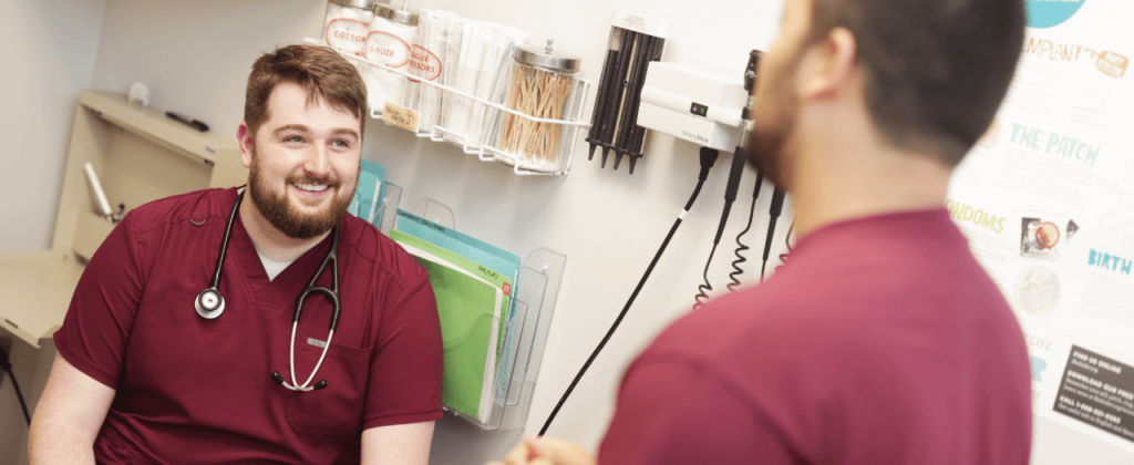 A male student in maroon scrubs with a stethoscope draped on his neck sits in a clinic setting and speaks with another male student, standing.